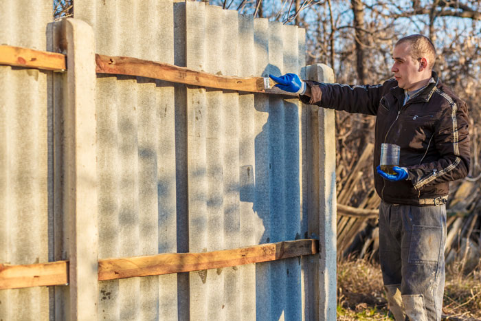 Man inspecting newly replaced fence by neighbor, standing outdoors with paintbrush and wearing blue gloves. Man inspecting newly replaced fence by neighbor, standing outdoors with paintbrush and wearing blue gloves.