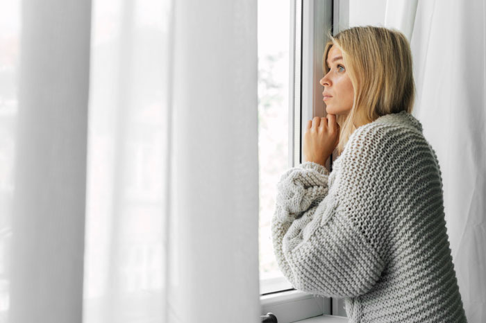 Woman in a gray sweater looking out the window at morning light, reflecting on neighbor drama and car panic alarm revenge.
