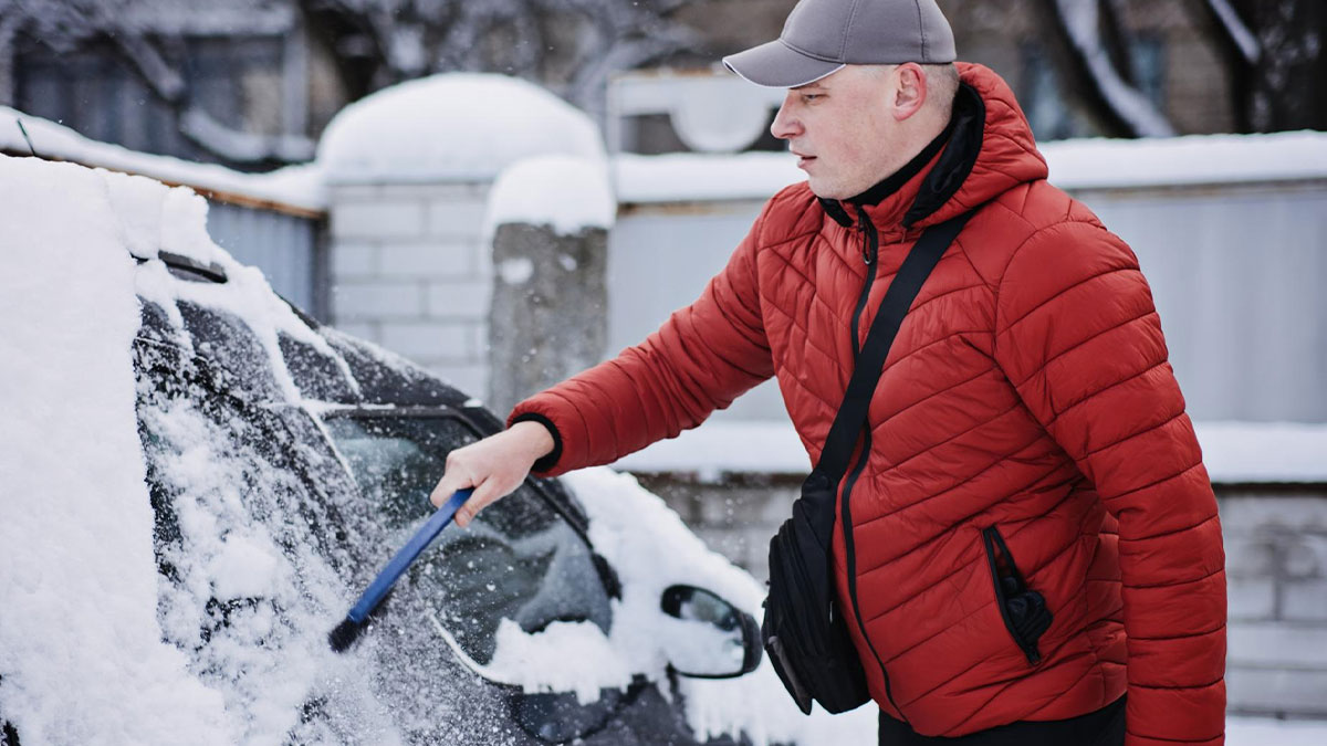 Man in a red jacket clearing snow from car window early morning, neighbor comes over to help couple at 6:30 AM.