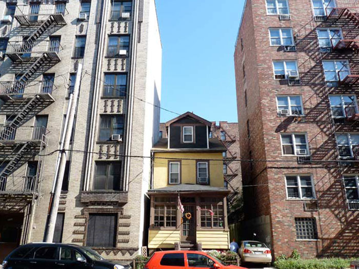 Small yellow house framed by tall concrete and brick apartment buildings with fire escapes and parked cars below.