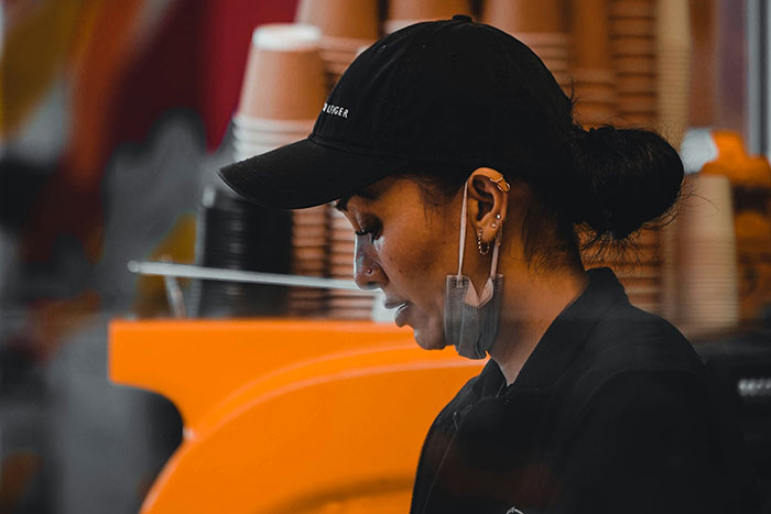 Gas station worker wearing black cap and mask under chin, focused on task behind counter with orange background.