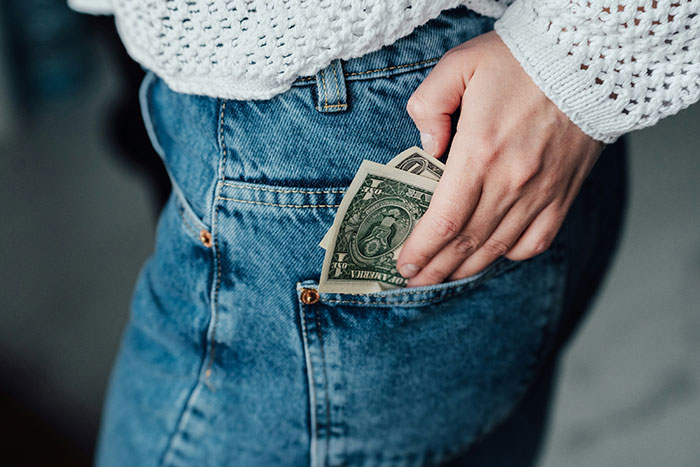 Hand putting dollar bills into the front pocket of blue jeans, representing gas station workers' stories and tips.