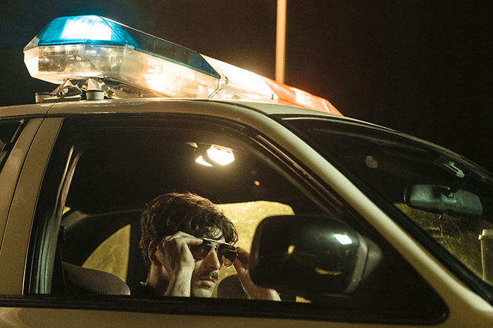 Gas station worker wearing sunglasses inside a police car at night sharing wild and unexpected stories.