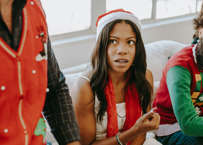Woman in a Santa hat looking confused during family Christmas traditions with others wearing festive sweaters nearby.