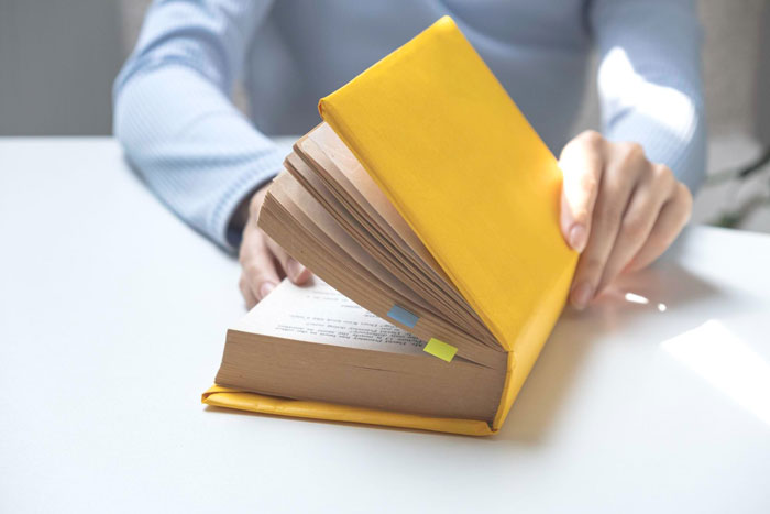 Person flipping through a yellow book carefully, symbolizing strict adherence to rules by the book in an office setting. Person flipping through a yellow book carefully, symbolizing strict adherence to rules by the book in an office setting.