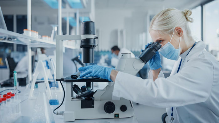 Scientist in lab coat and mask using microscope in a busy laboratory illustrating strict office rules by the book. Scientist in lab coat and mask using microscope in a busy laboratory illustrating strict office rules by the book.