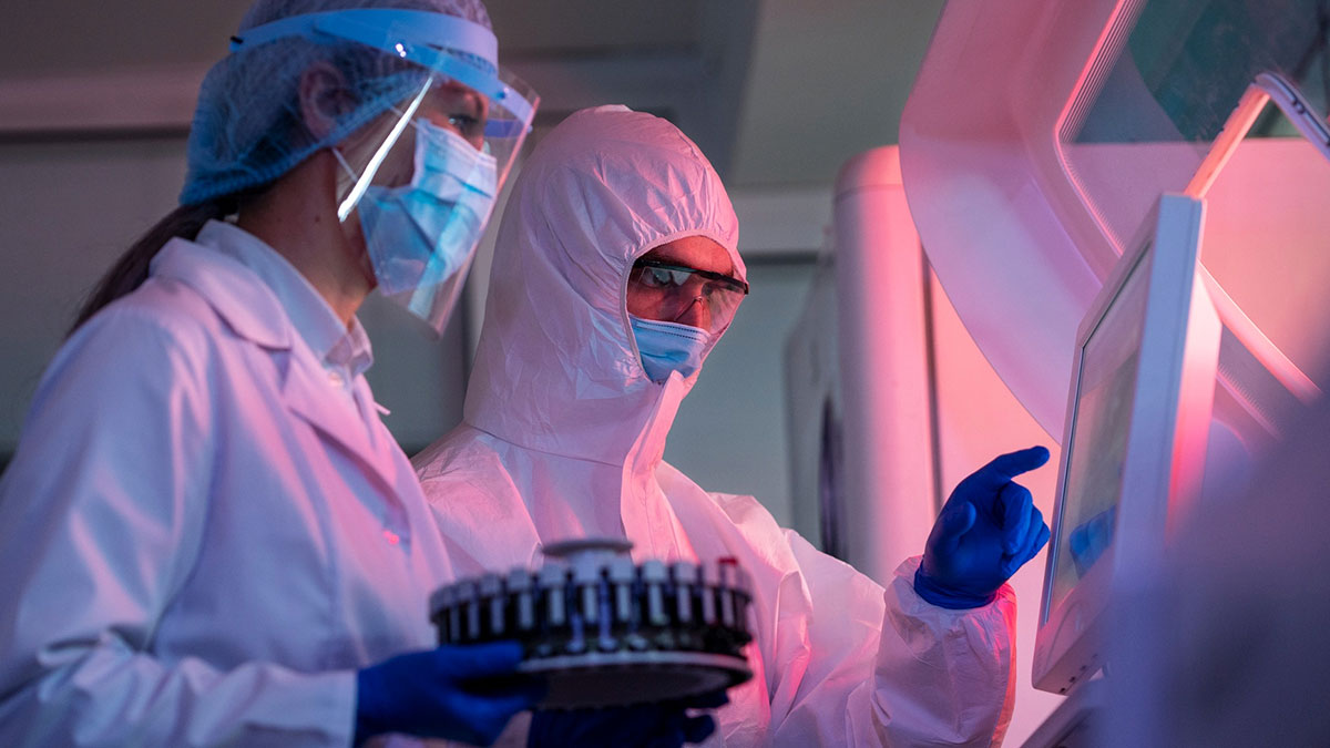 Two lab workers in protective gear and masks operating lab equipment, emphasizing strict procedures done by the book.