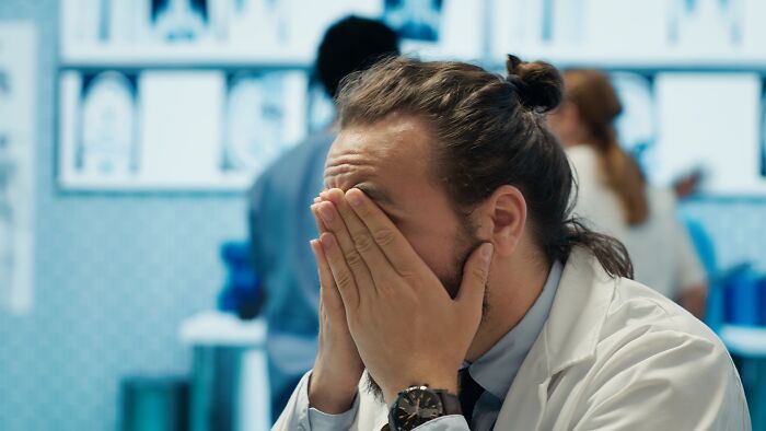 Doctor in white coat covering face with hands, appearing stressed or overwhelmed in a busy medical setting, creepy cases concept