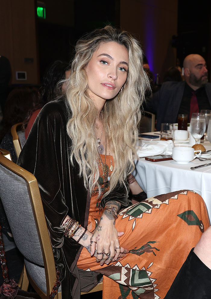Paris Jackson seated at an event, wearing a patterned dress and showcasing her natural face without visible deformities from substance use. Paris Jackson seated at an event, wearing a patterned dress and showcasing her natural face without visible deformities from substance use.
