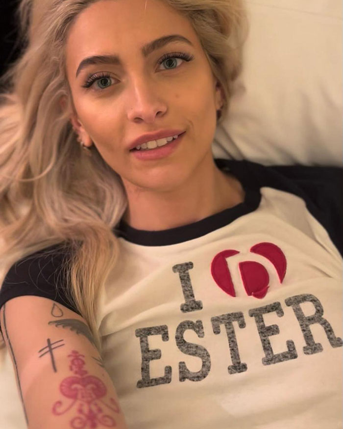 Paris Jackson lying down with tattoos on arm, smiling and wearing a white shirt with black sleeves. Paris Jackson lying down with tattoos on arm, smiling and wearing a white shirt with black sleeves.