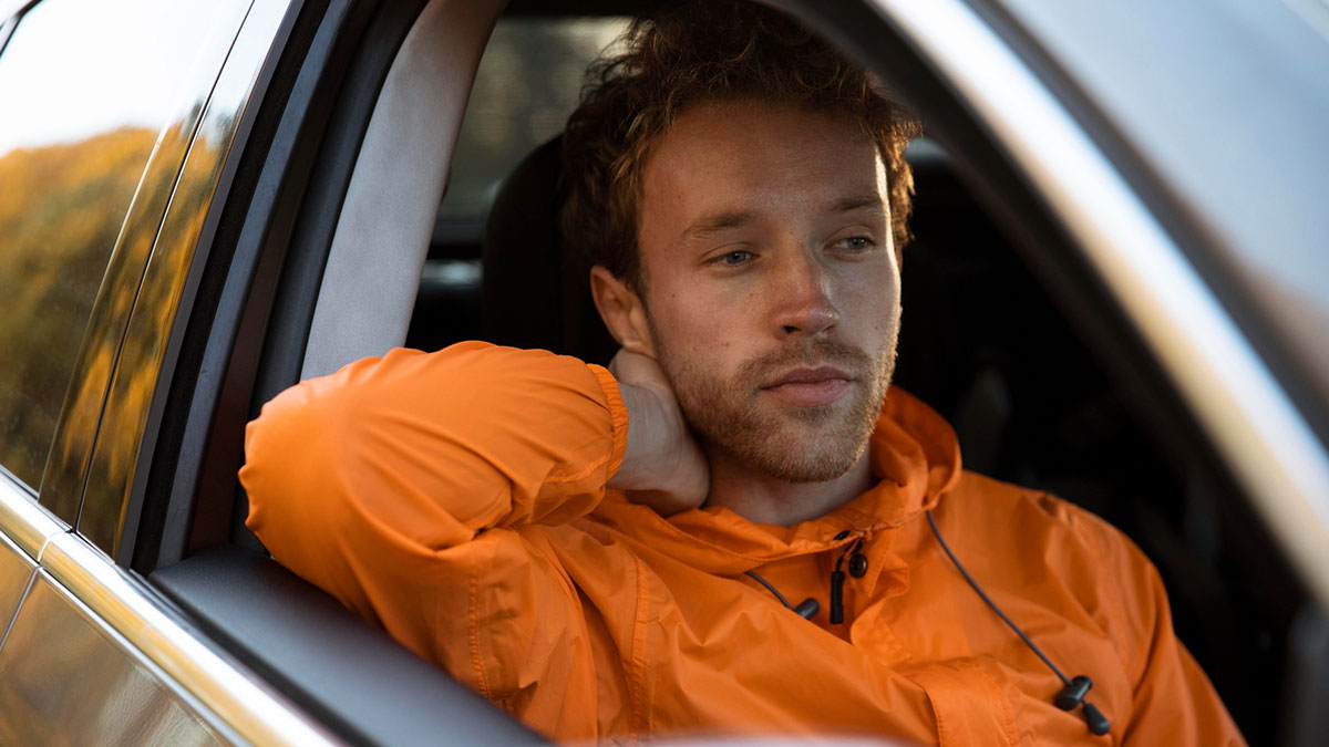 Young man in an orange jacket sitting inside a car, reflecting on disappeared people starting new lives experiences.