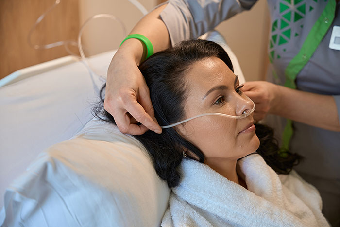 Woman with nasal oxygen tube resting in hospital bed while nurse adjusts the tubing, representing coma survivors' recovery stories.