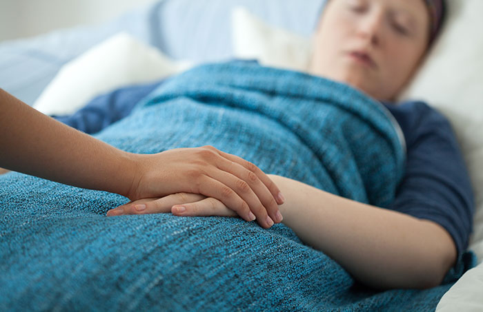 Patient covered with a blue blanket lying in bed while holding the hand of a visitor in a coma survivor story context.