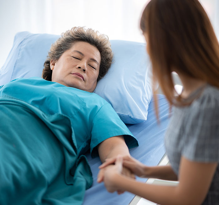 Older woman in hospital bed holding hands with visitor, illustrating coma survivors sharing their recovery stories.