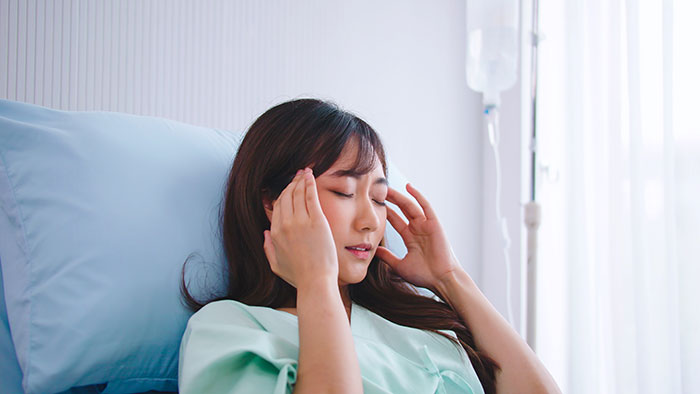 Young woman in hospital gown resting in bed, appearing pensive, representing coma survivors sharing their stories.