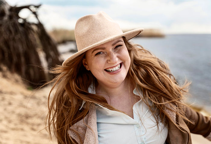 Woman with long hair wearing a hat smiling on a beach, representing people who walked away from their old lives.