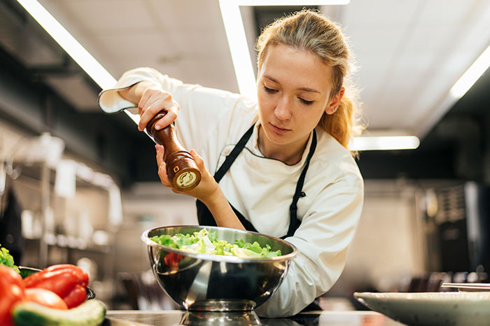 Young woman who walked away from her old life seasoning salad in a professional kitchen setting