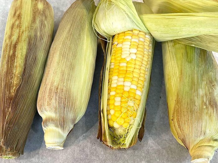 Four ears of corn with husks, one partially peeled to reveal yellow and white kernels, illustrating kitchen tips for cooking. Four ears of corn with husks, one partially peeled to reveal yellow and white kernels, illustrating kitchen tips for cooking.