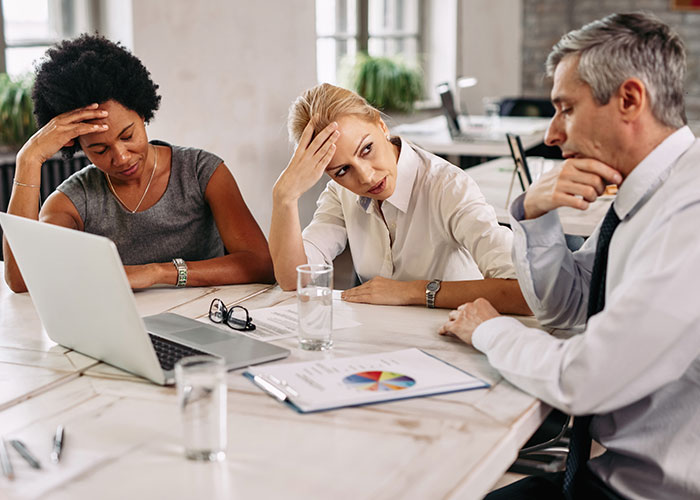 Three stressed businesspeople at a meeting table with laptop and documents, dealing with package delivery issues.