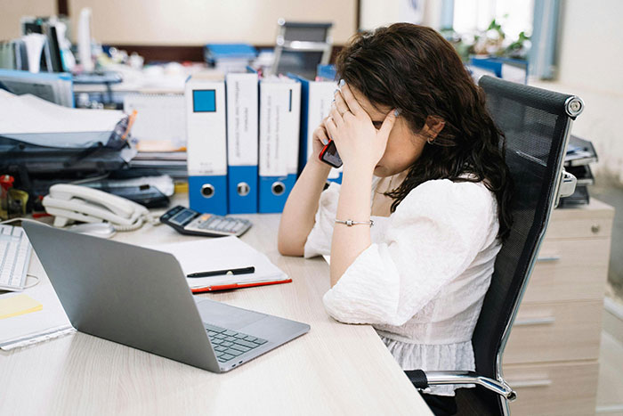 Woman frustrated at office desk, holding phone and covering face, illustrating coworker dispute over expensive jacket. Woman frustrated at office desk, holding phone and covering face, illustrating coworker dispute over expensive jacket.