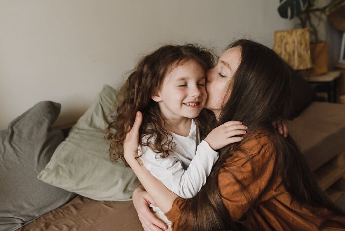 Mother hugging and kissing her smiling child on the couch, capturing moments of childhood habits and unexpected normalcy.