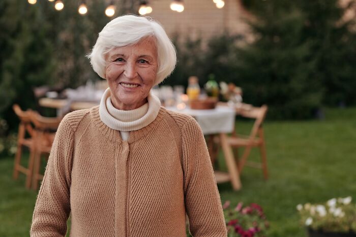 Smiling elderly woman outdoors in cozy sweater, with blurred garden and string lights in the background.