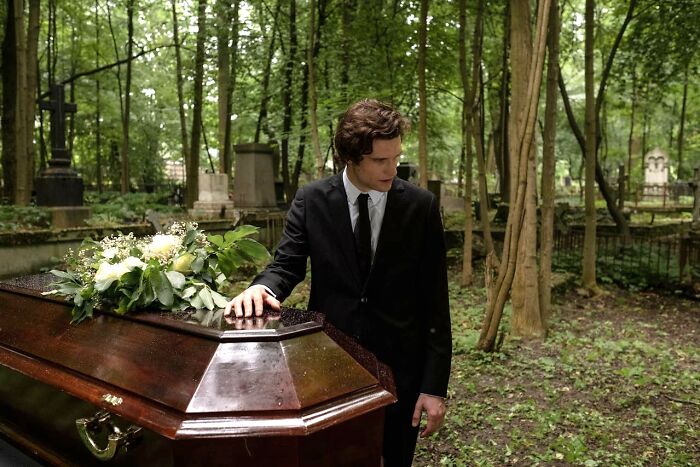 Man in black suit standing beside a coffin outdoors surrounded by trees, evoking uniquely awkward wedding moments.