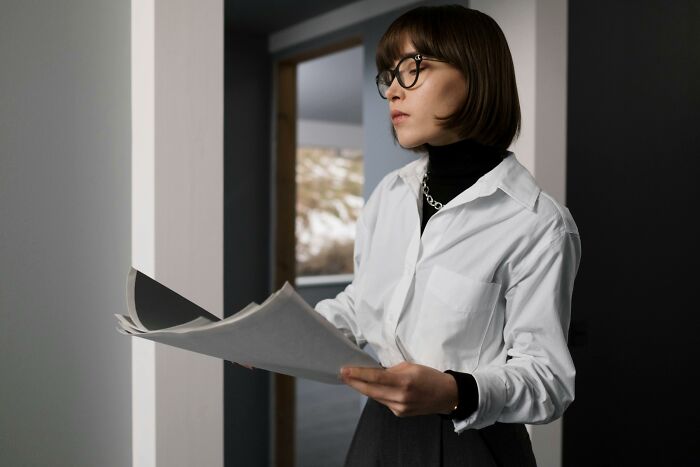 Woman in glasses and white shirt holding papers, reflecting on infuriating work situations involving old habits.