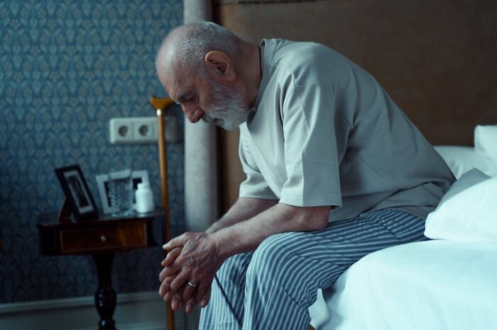 Elderly man sitting on bed, deep in thought, reflecting on heavy confessions he refused to take to his grave.