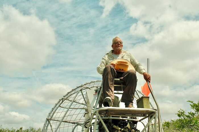 Older man sitting on an airboat holding a hat, surrounded by nature under a cloudy sky in a bizarre moment.