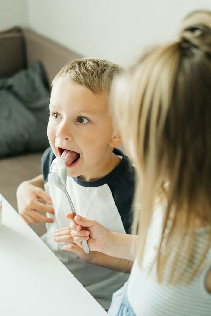 Young boy displaying unusual childhood habits while a girl feeds him with a spoon at a table indoors.