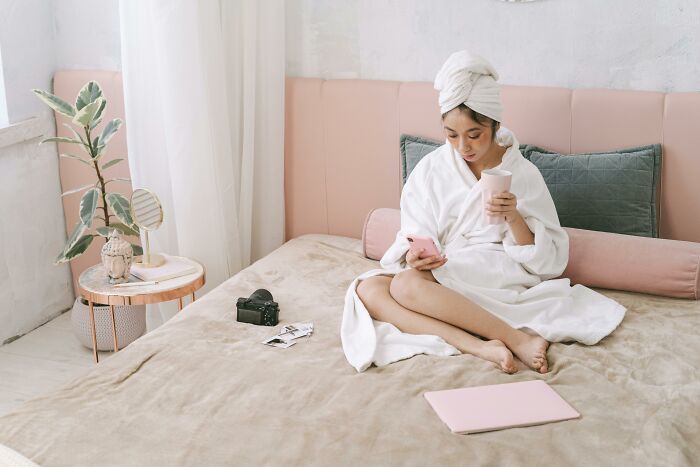 Woman in bathrobe and towel on head, relaxing on bed with coffee and phone, showing relatable everyday moments.