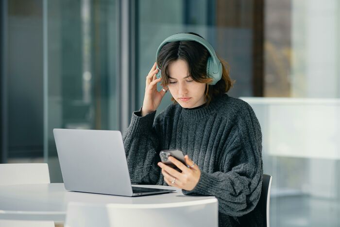 Young woman wearing headphones, using smartphone and laptop, relatable to ridiculous but relatable things people do.