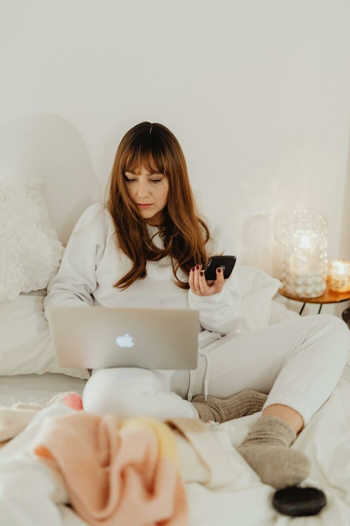 Woman in casual white clothes sitting on bed, using laptop and phone, reflecting on childhood habits not normal experiences.