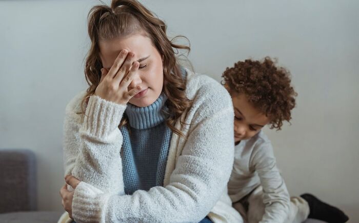 Stressed mother sitting with hand on face while child behind her, capturing funny and serious moments parents realize raising monsters.