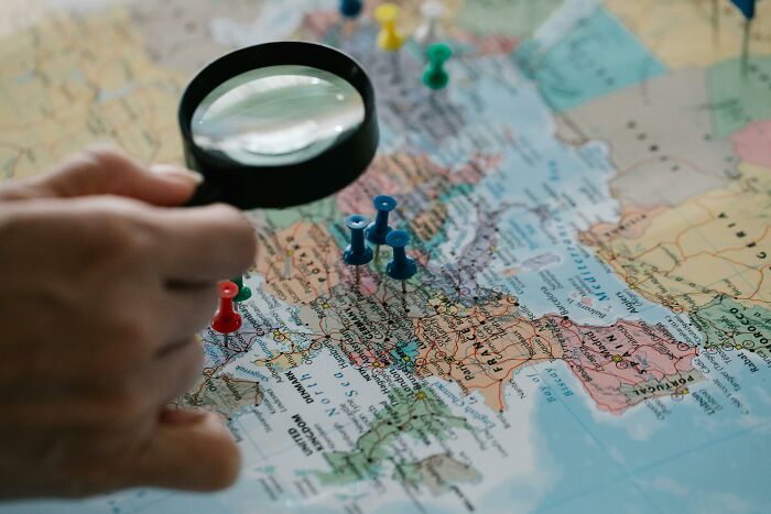 Hand holding magnifying glass over a world map with pins, highlighting geography questions to test knowledge.