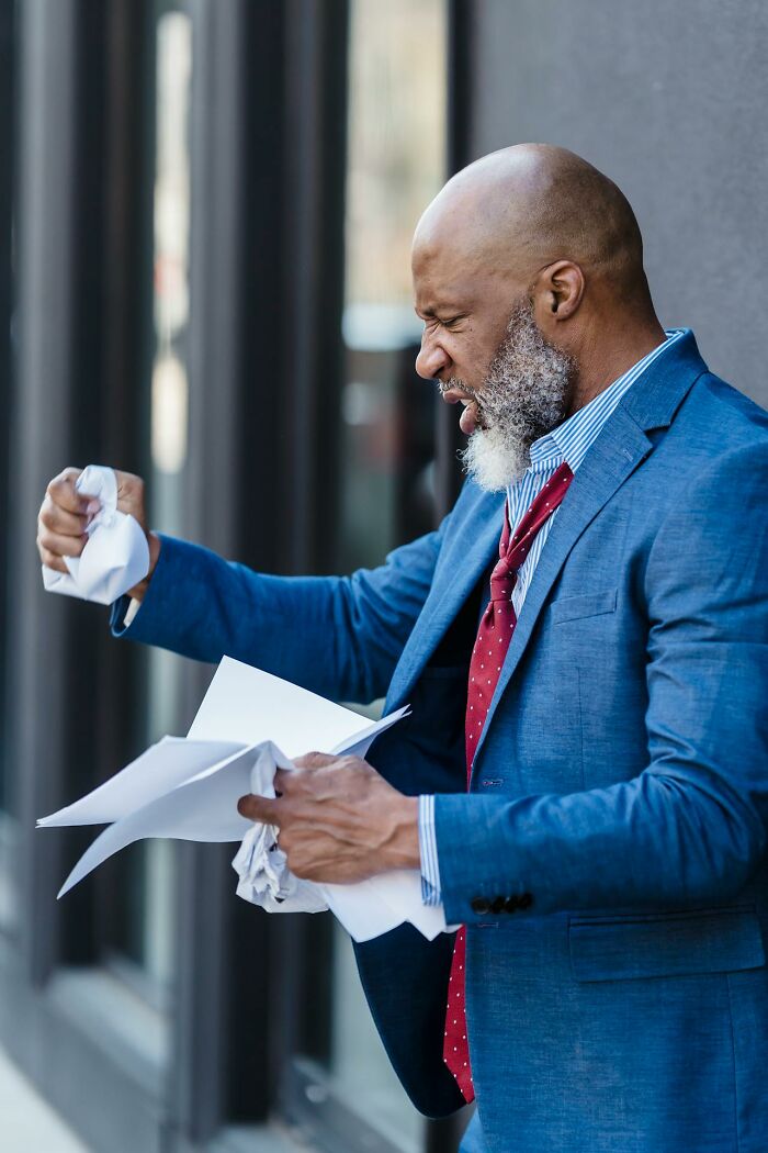 Angry man in blue suit crumpling papers outside building, showing intense frustration and stress like crazy Karens. Angry man in blue suit crumpling papers outside building, showing intense frustration and stress like crazy Karens.