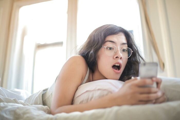 Young woman with glasses lying on bed, looking shocked at phone screen, illustrating relatable and ridiculous habits.