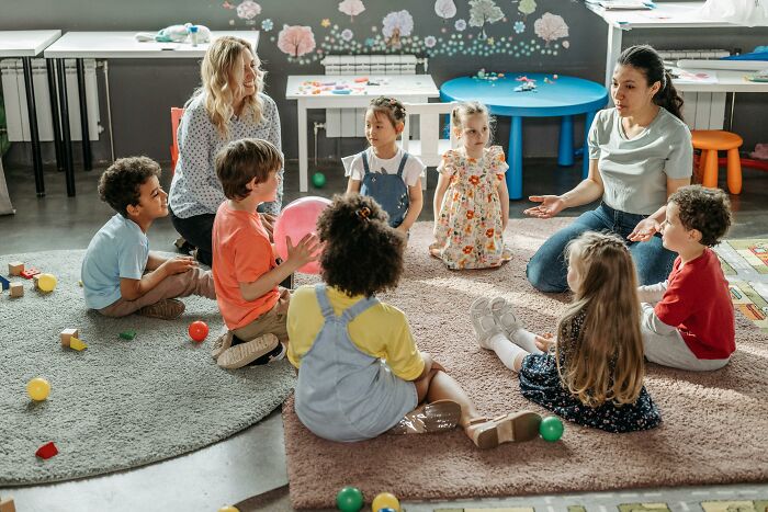 Teacher sitting on rug with children in a classroom, illustrating infuriating examples experienced at work.