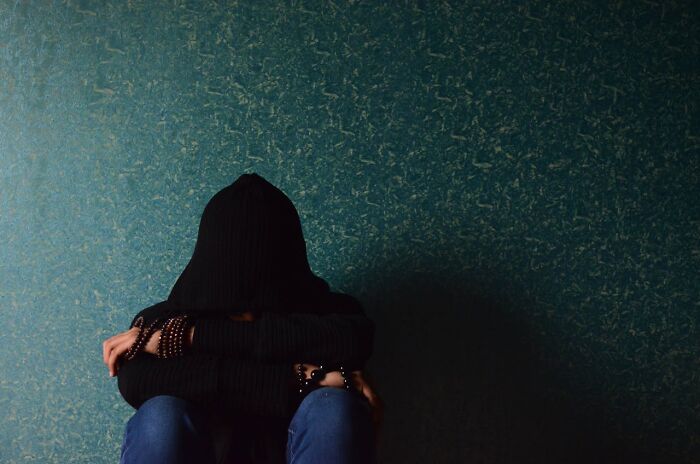 Person wearing a black hoodie sitting against textured wall, conveying a bizarre moment of isolation and confusion.