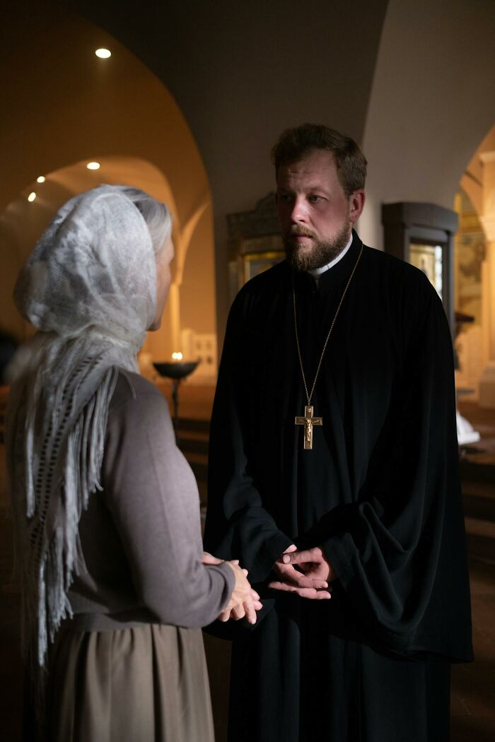 A priest wearing a cross talks with a woman inside a dimly lit church, symbolizing heavy confessions and secrecy.