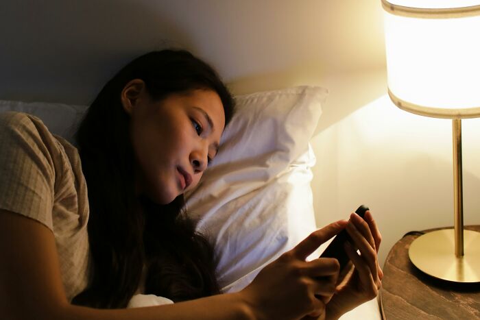 Young woman lying in bed using smartphone at night, illustrating relatable and ridiculous everyday behaviors.