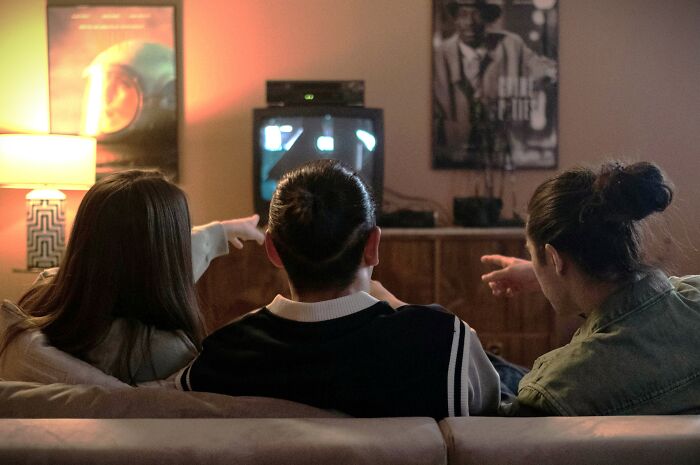 Three people sitting on a couch pointing at a TV screen, enjoying iconic TV show opening credits together. Three people sitting on a couch pointing at a TV screen, enjoying iconic TV show opening credits together.