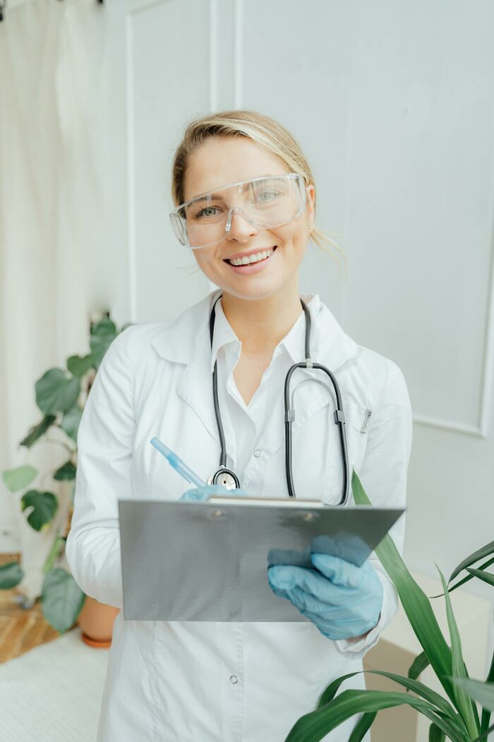 Psych ward nurse wearing safety glasses and gloves, holding clipboard and pen, smiling in clinical setting.