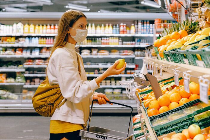 Woman wearing mask shopping for fruit in grocery store, illustrating relatable and ridiculous everyday habits people are guilty of.
