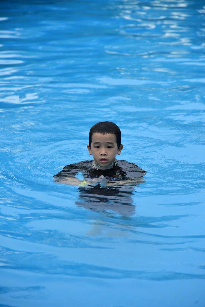 Young boy in black shirt swimming alone in a pool, capturing a bizarre moment of solitude and reflection.