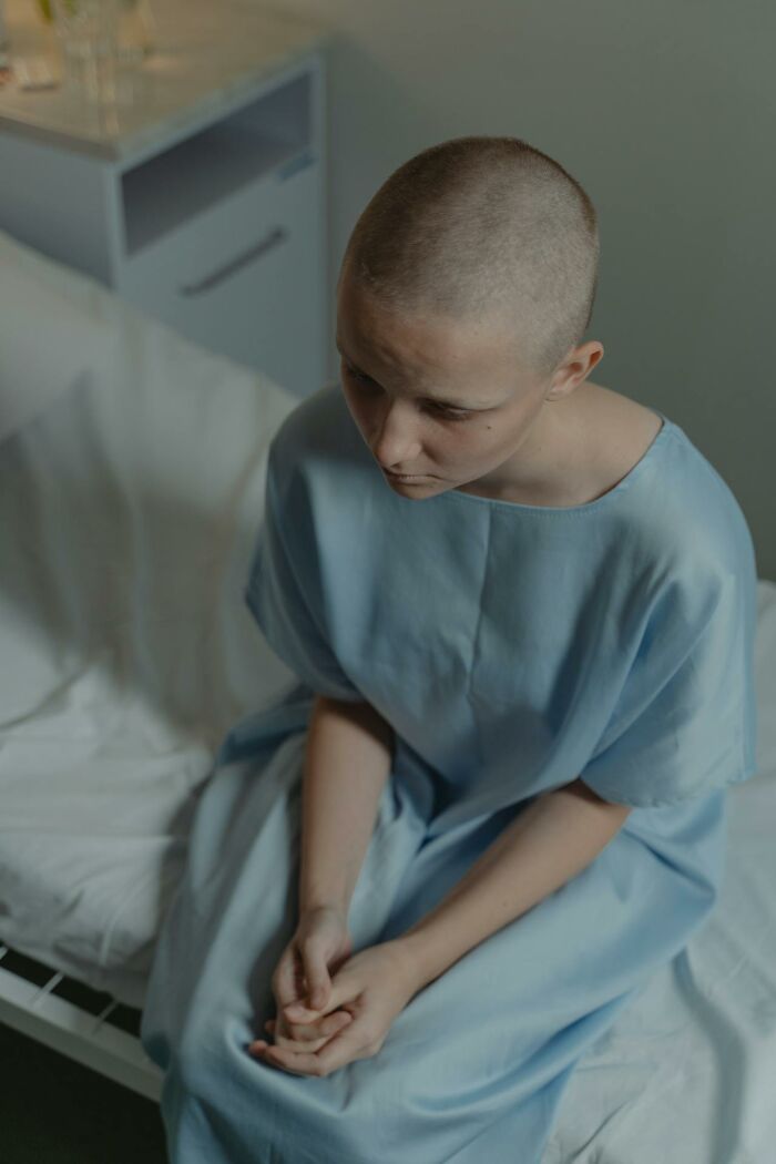 Person with shaved head wearing hospital gown sitting on bed, reflecting on experiences of dying and coming back to life.