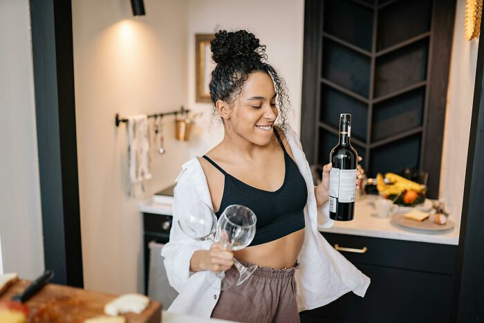 Young woman in casual clothes holding a wine bottle and glasses, enjoying a relatable and ridiculous moment at home.