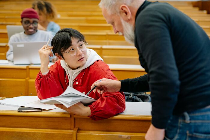 Student in red jacket looking confused while teacher explains notes in lecture hall showing signs child never loved properly in education context