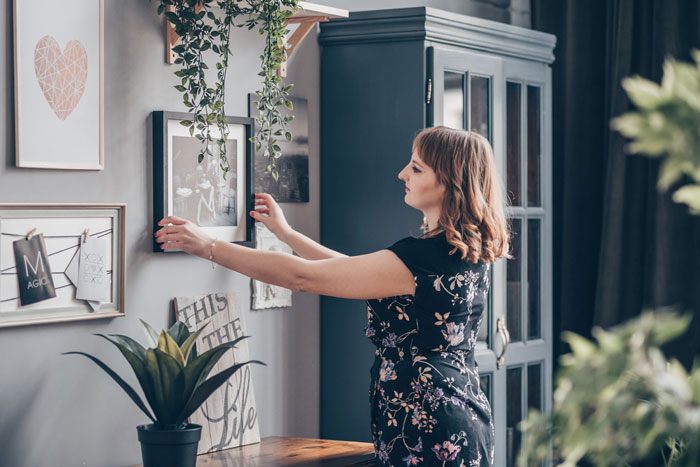 Woman adjusting framed photos on wall in a home, illustrating jealous wife reacting to hubby’s mom having pics of his ex.